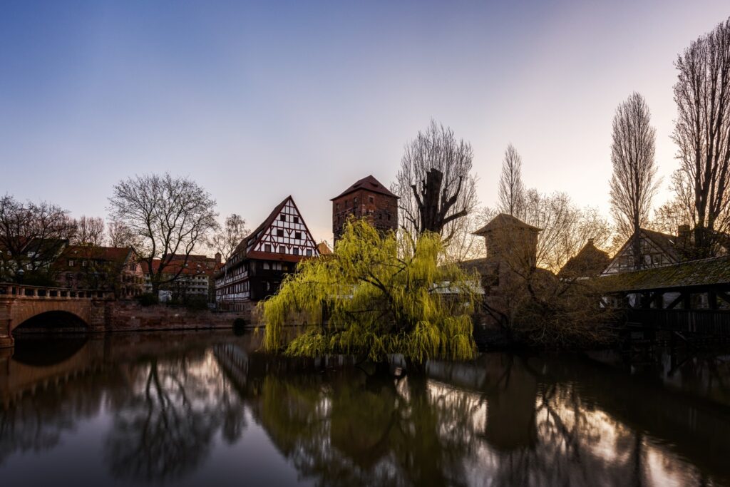 Historische Fachwerkhäuser am Fluss, Brücke, Weiden, Turm – malerische Altstadtansicht bei Sonnenuntergang.