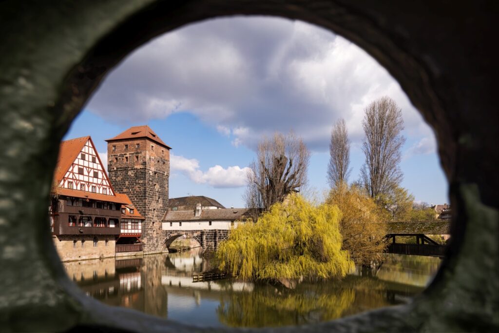 Durch eine runde, dunkle Öffnung ist eine historische Flusslandschaft mit einem Fachwerkhaus, einem Turm, einer Steinbrücke und Bäumen zu sehen, deren Spiegelungen sich im Wasser unter einem teilweise bewölkten Himmel zeigen.