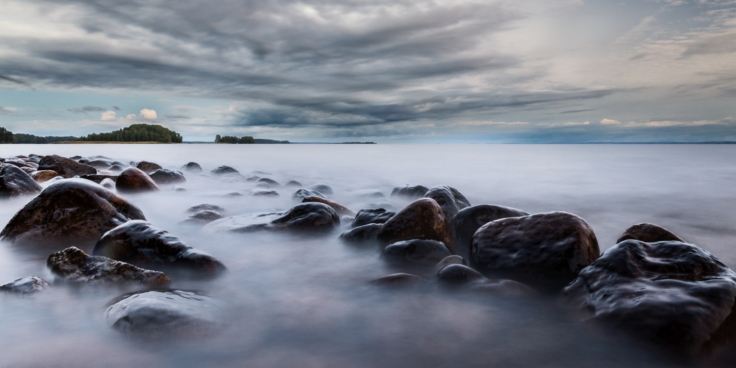 Langzeitbelichtete Uferlandschaft mit runden Steinen am Ufer, ruhiges Wasser und dramatischer Wolkenhimmel.