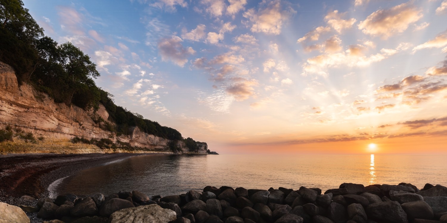 Küstenlandschaft: Kiesstrand, Felsklippen links, ruhiges Meer, Sonnenuntergang über dem Horizont.