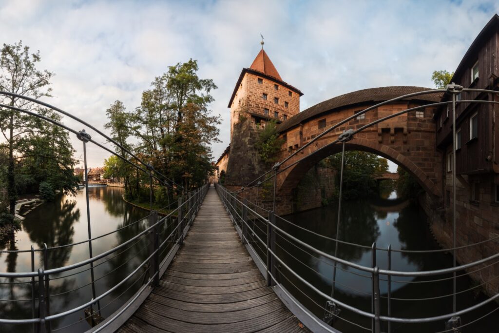 Historische Steinbrücke mit Turm über dem Fluss; Fußgänger-Holzsteg und malerische Uferlandschaft.