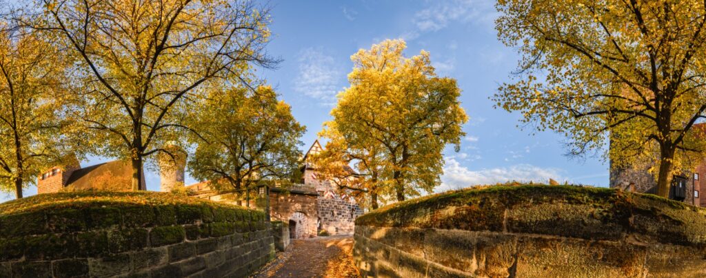 Herbstliche Burgmauer mit Tor, goldene Baumblätter, gepflasterter Weg – historische Burganlage unter blauem Himmel.