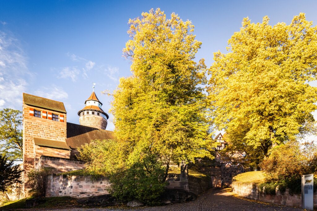 Historische Festungsanlage mit Turm, Kopfsteinpflasterweg und goldenem Herbstlaub vor blauem Himmel