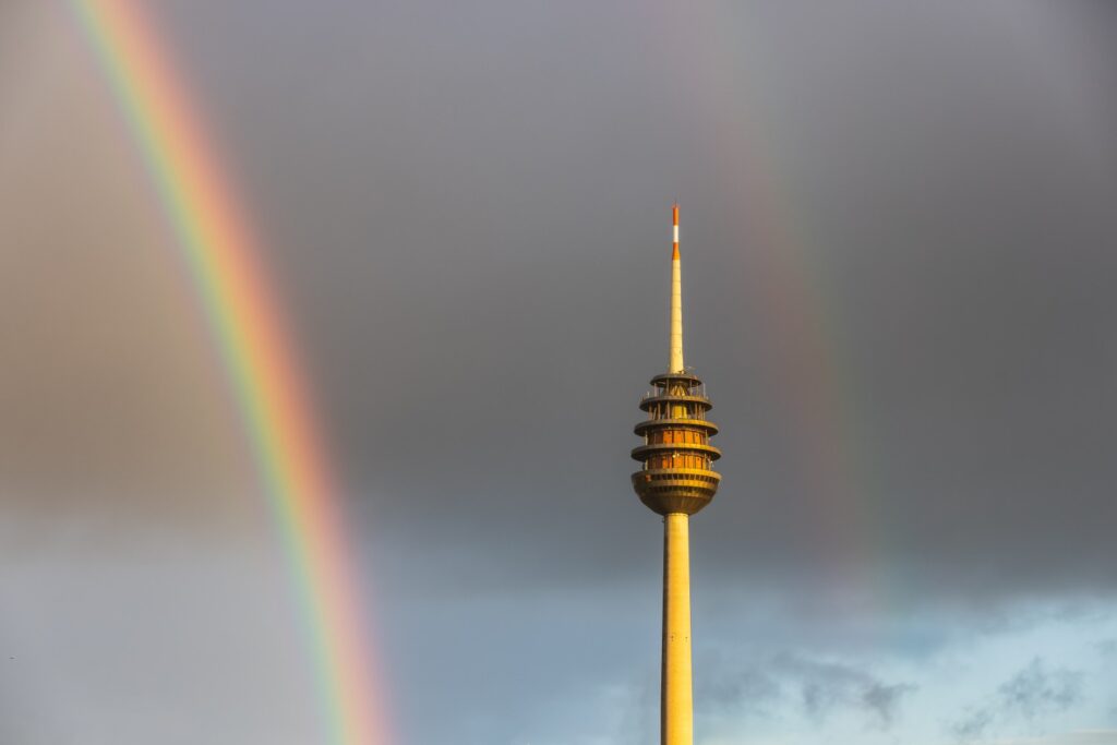 Fernsehturm mit doppeltem Regenbogen vor grauem Himmel.