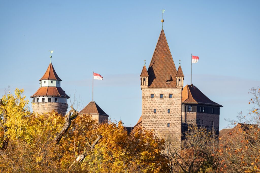 Historische Burganlage mit roten Ziegeldächern, Türmen und Flaggen, umgeben von Herbstlaub vor blauem Himmel.