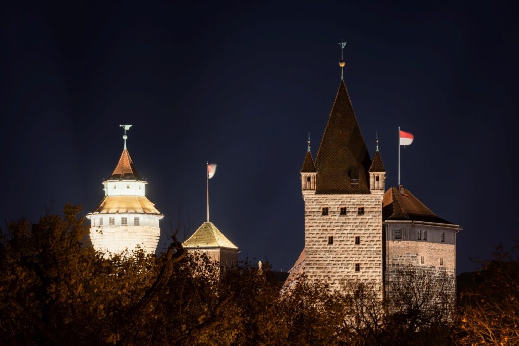 Die Aufnahme zeigt eine bei Nacht hell erleuchtete Burganlage mit markanten Türmen, über denen rot weiße Flaggen wehen, und dunklem Blattwerk im Vordergrund.