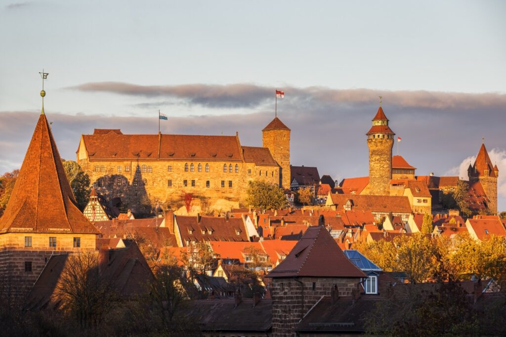 Eine imposante Burganlage mit mehreren Türmen überragt eine darunter liegende Stadt mit zahlreichen Häusern und roten Dächern, alles beleuchtet vom warmen Licht des späten Tages unter einem bewölkten Himmel.
