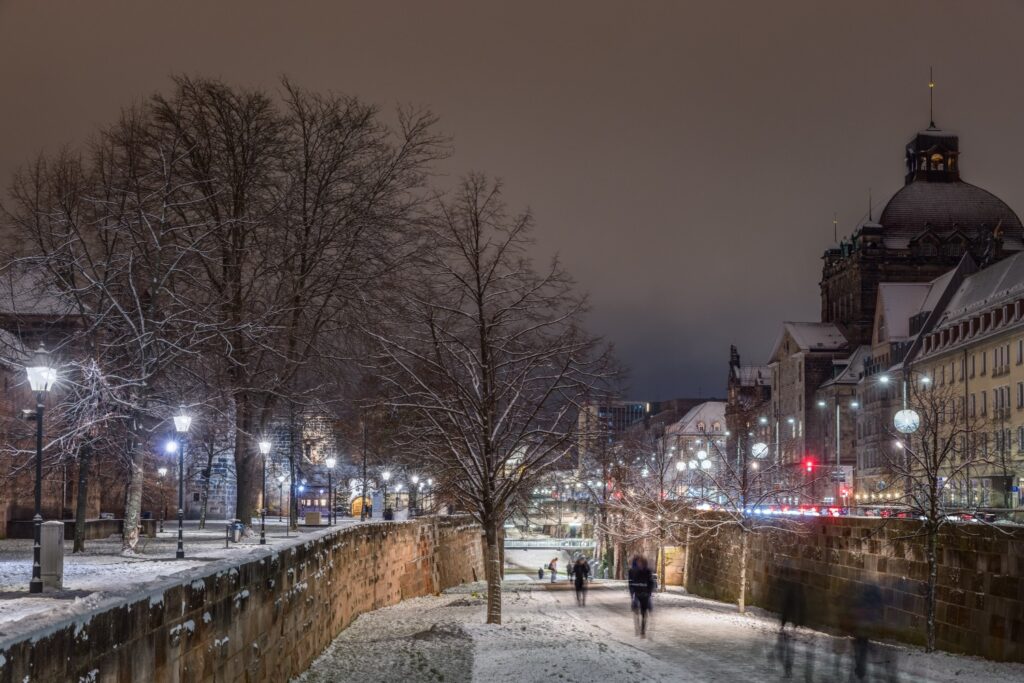 Verschneite Uferpromenade in der Nacht mit Laternen, historischen Gebäuden und Kuppel, Fußgänger und Radfahrer unterwegs.