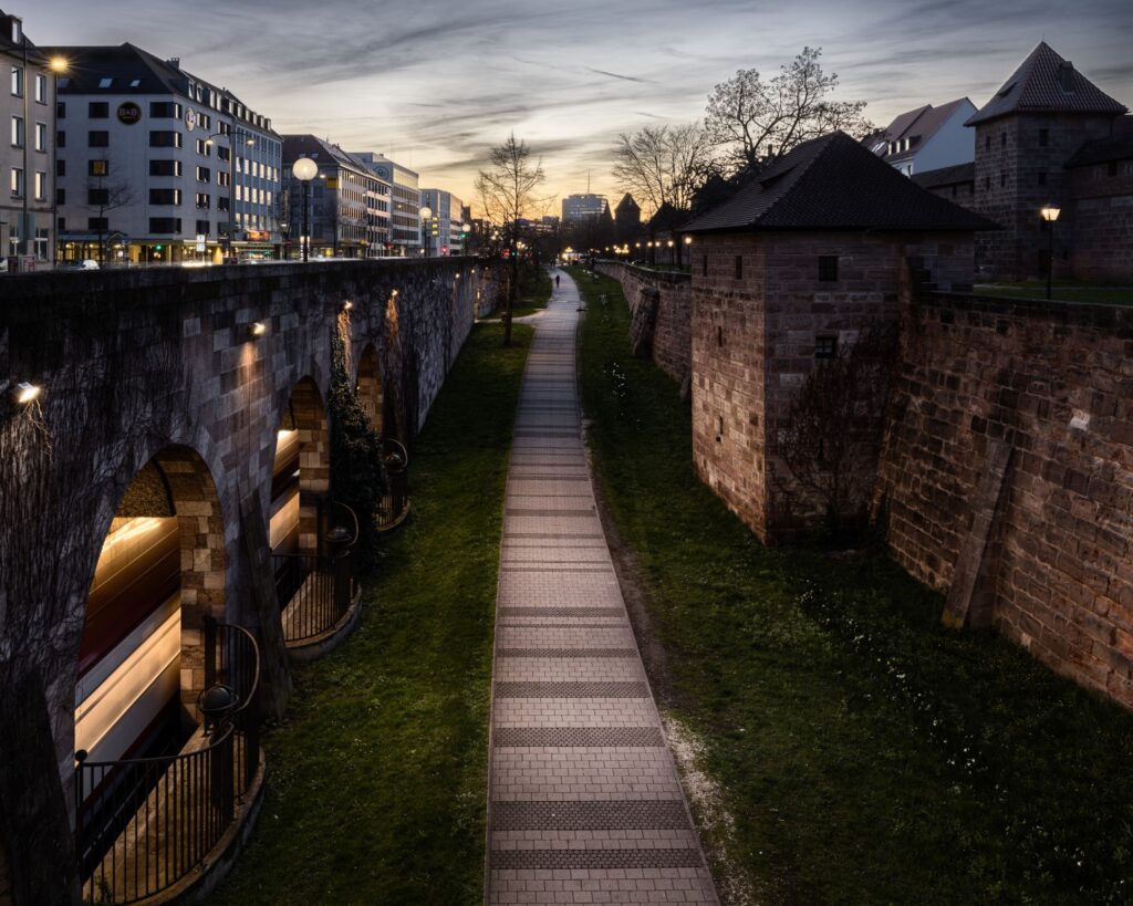 Abendlicher Blick auf historische Stadtmauer mit Arkaden und begehbarer Promenade zwischen Mauern und moderner Innenstadt.