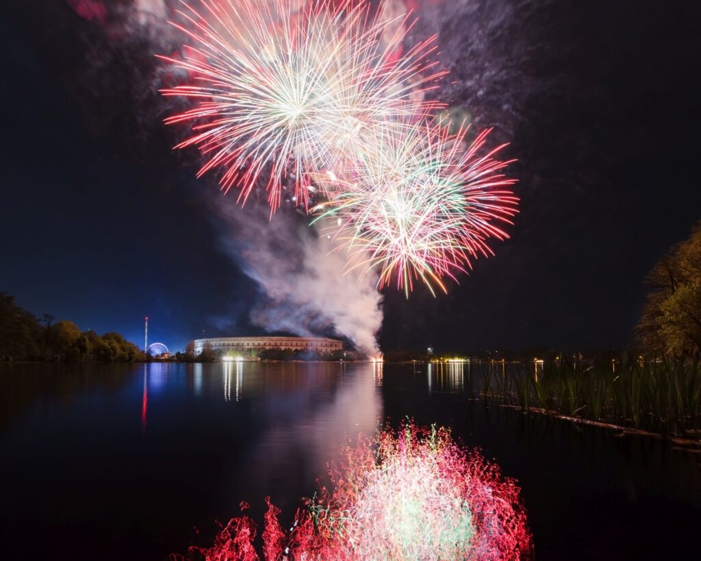 Man sieht ein farbenprächtiges Feuerwerk am Nachthimmel, dessen Lichter sich im ruhigen Wasser eines Sees spiegeln, an dessen Ufer ein großes Gebäude und ein Riesenrad beleuchtet sind.