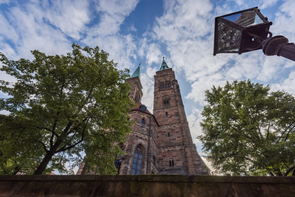 Historische Kirche mit zwei Türmen und grünen Spitzen, von Bäumen umgeben, unter blauem Himmel; Laterne im Vordergrund