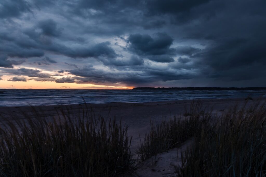 Sturm am Strand bei Abenddämmerung: dunkle Wolken, orangefarbener Horizont, brechende Wellen, Dünengras im Vordergrund.
