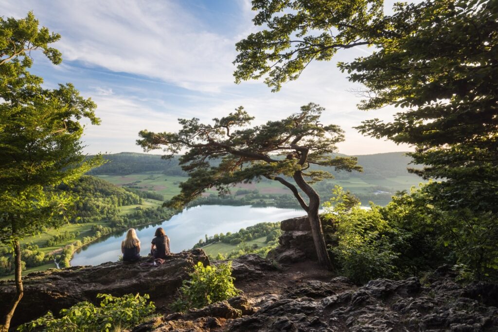 Zwei Personen sitzen auf einer Felsklippe und genießen den Panoramablick über Fluss, Wälder und grüne Hügellandschaft.