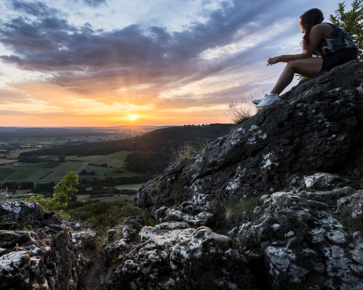 Person sitzt auf einem Felsen und beobachtet den Sonnenuntergang über einer grünen Landschaft, mit Wäldern und Feldern im Hintergrund. Die Szene vermittelt ein Gefühl von Ruhe und Naturverbundenheit.