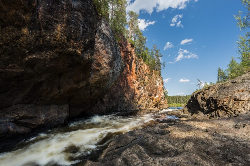Felsen und Wasserströmung in einer malerischen Landschaft mit Bäumen und blauem Himmel, ideal für Naturfotografie und Outdoor-Aktivitäten.
