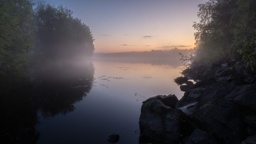 Ruhige Morgenstimmung am See mit Nebel, umgeben von Bäumen und Felsen, während die Sonne aufgeht und den Himmel in sanften Farben erleuchtet.