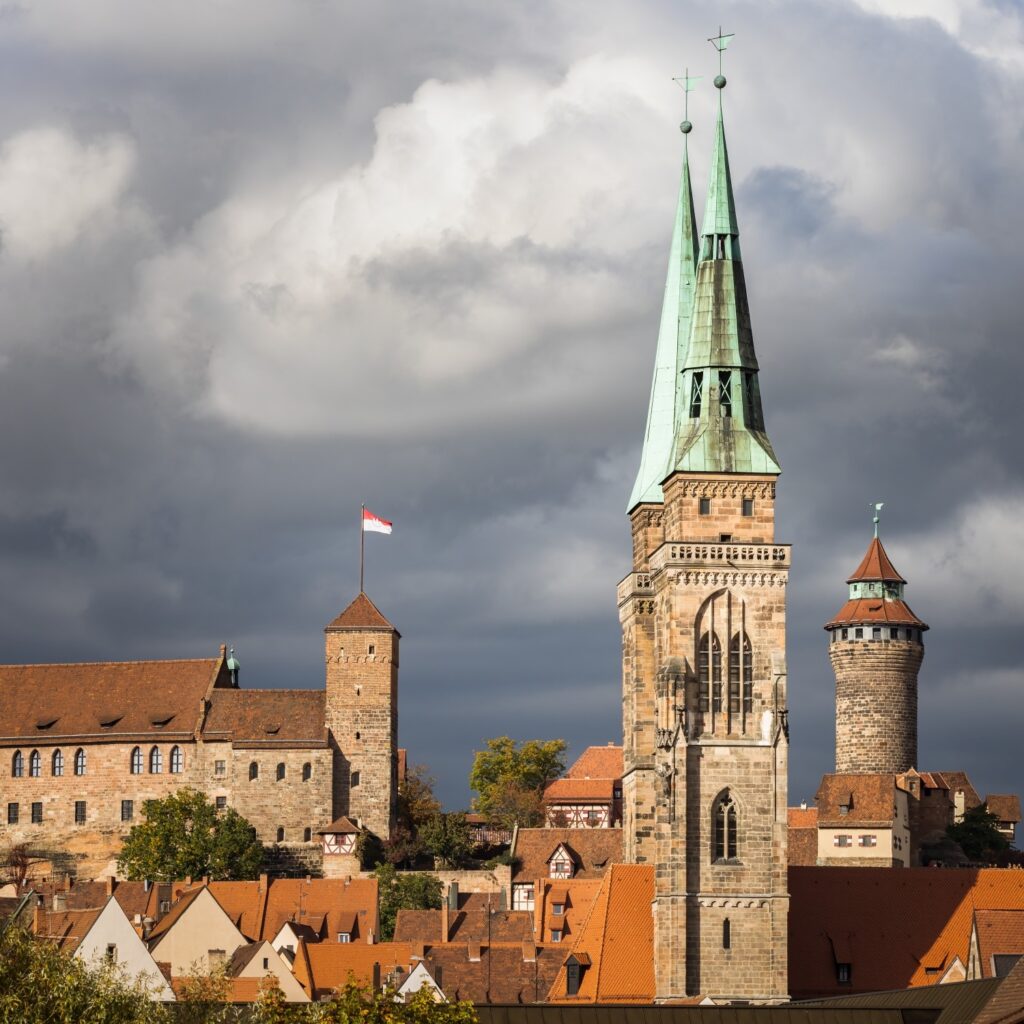 Eine historische Stadtansicht zeigt eine Kirche mit zwei markanten grünen Turmspitzen, eine Burg mit wehender rotweißer Fahne und zahlreiche rote Dächer unter einem dramatischen Wolkenhimmel.