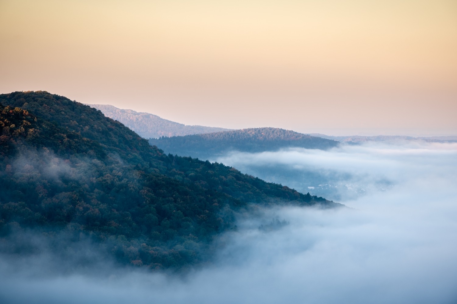 Morgennebel über bewaldeten Bergen, sanfte Hügelkette im Dunst, warmer Sonnenaufgang am Horizont.