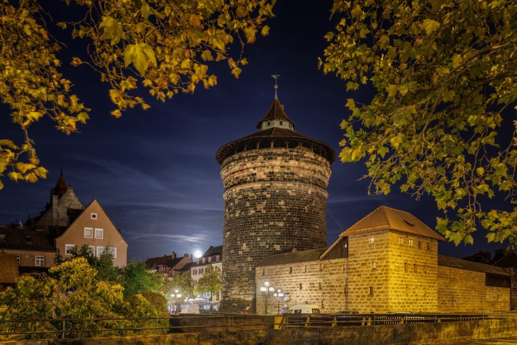 Ein großer runder Steinturm ragt nachts unter einem mondhellen Himmel empor, eingerahmt von herbstlichem Laub und umgeben von beleuchteten Gebäuden.