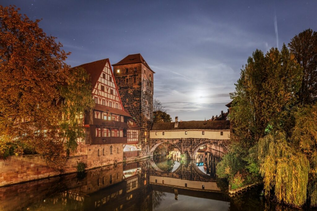 Das Bild zeigt eine nächtliche Szene mit historischen Fachwerkhäusern und einem Steinturm, die sich zusammen mit einer Bogenbrücke in einem Fluss spiegeln, umrahmt von herbstlichen Bäumen unter einem mondhellen Himmel mit Sternen.