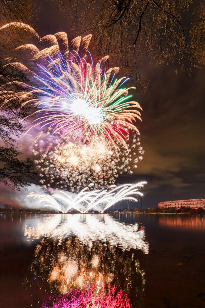Ein vielfältiges Feuerwerk erhellt den dunklen Nachthimmel über einem See, wobei sich die bunten Explosionen und ein Gebäude am gegenüberliegenden Ufer im Wasser spiegeln, eingerahmt von kahlen Baumästen.