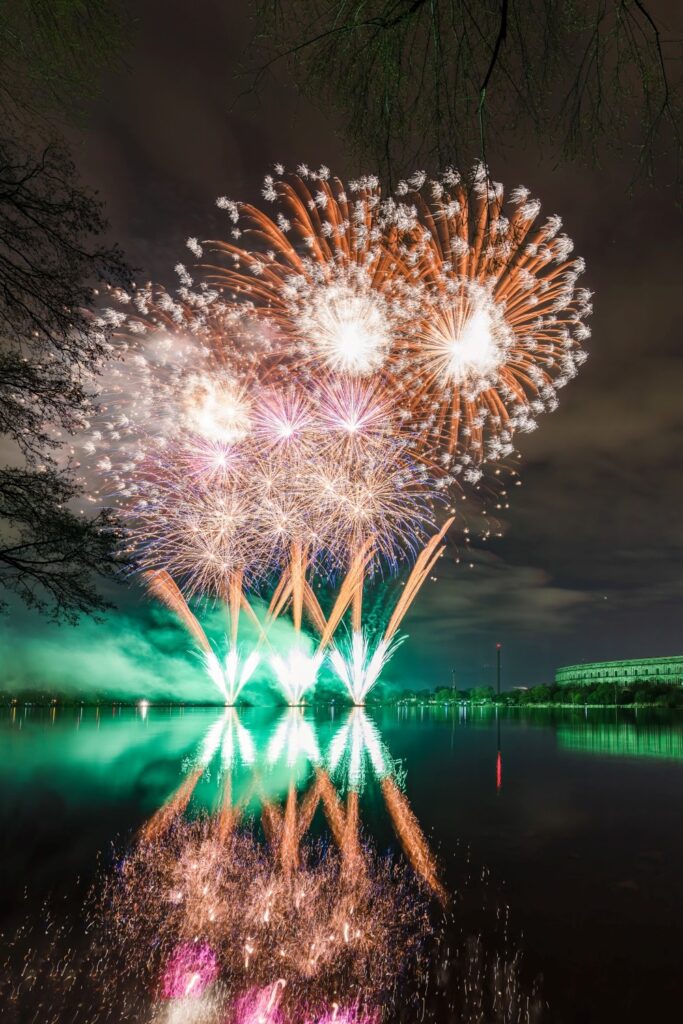 Ein spektakuläres Feuerwerk erhellt den Nachthimmel über einem Gewässer, wobei die bunten Explosionen und grünen Lichteffekte zusammen mit einem weit entfernten Gebäude und Bäumen im Wasser reflektiert werden.
