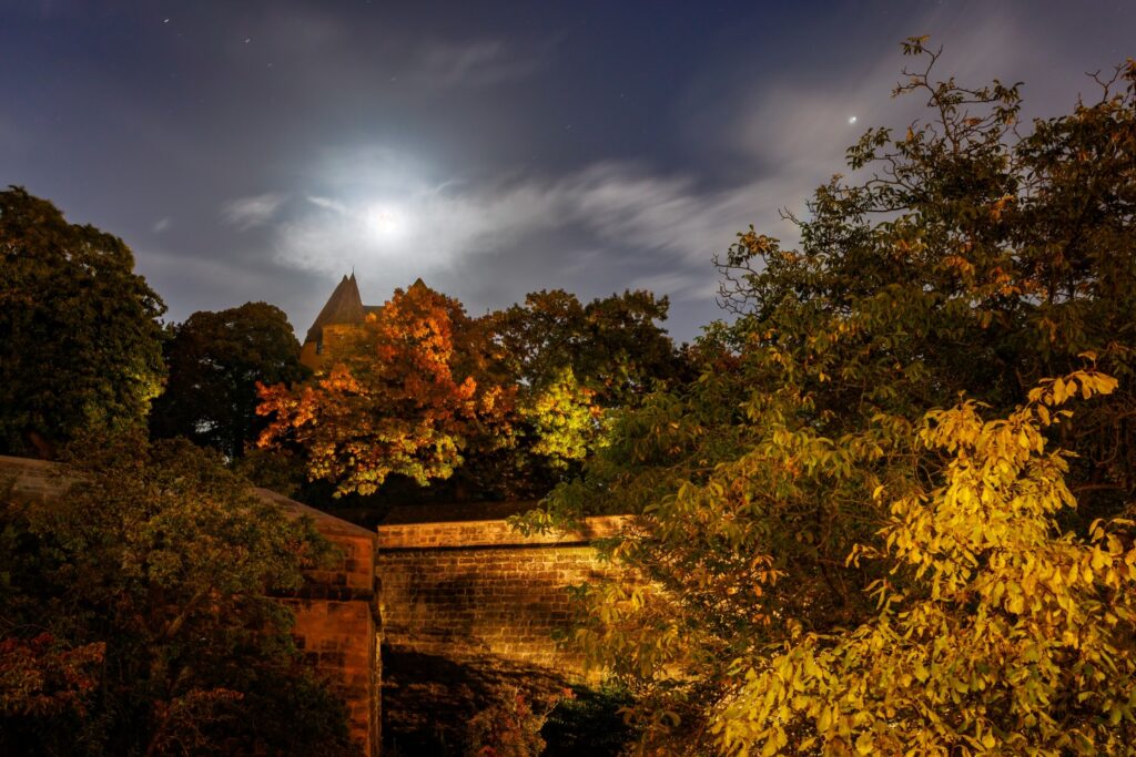 Nachtaufnahme einer alten Burganlage mit Mondschein, beleuchteten Herbstbäumen und Steinmauern im Vordergrund.