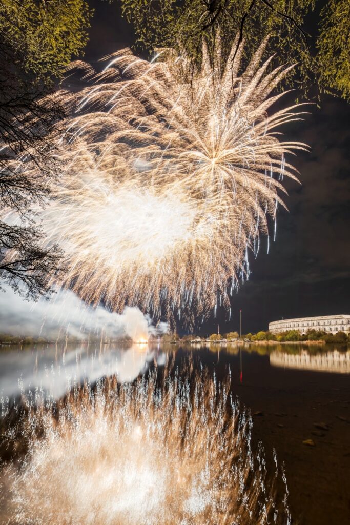 Ein spektakuläres Feuerwerk in strahlendem Gold und Weiß erhellt den Nachthimmel über einem See, dessen Oberfläche die explodierenden Lichter und ein großes Gebäude am Ufer widerspiegelt, während Äste und Blätter das obere Bildfeld umrahmen.