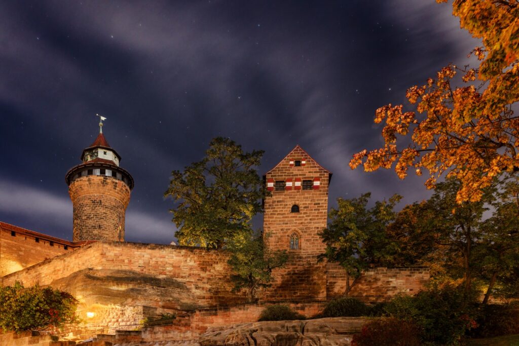 Nachtaufnahme einer historischen Burgmauer mit Rundturm und rechteckigem Turm, von Herbstlaub umgeben.