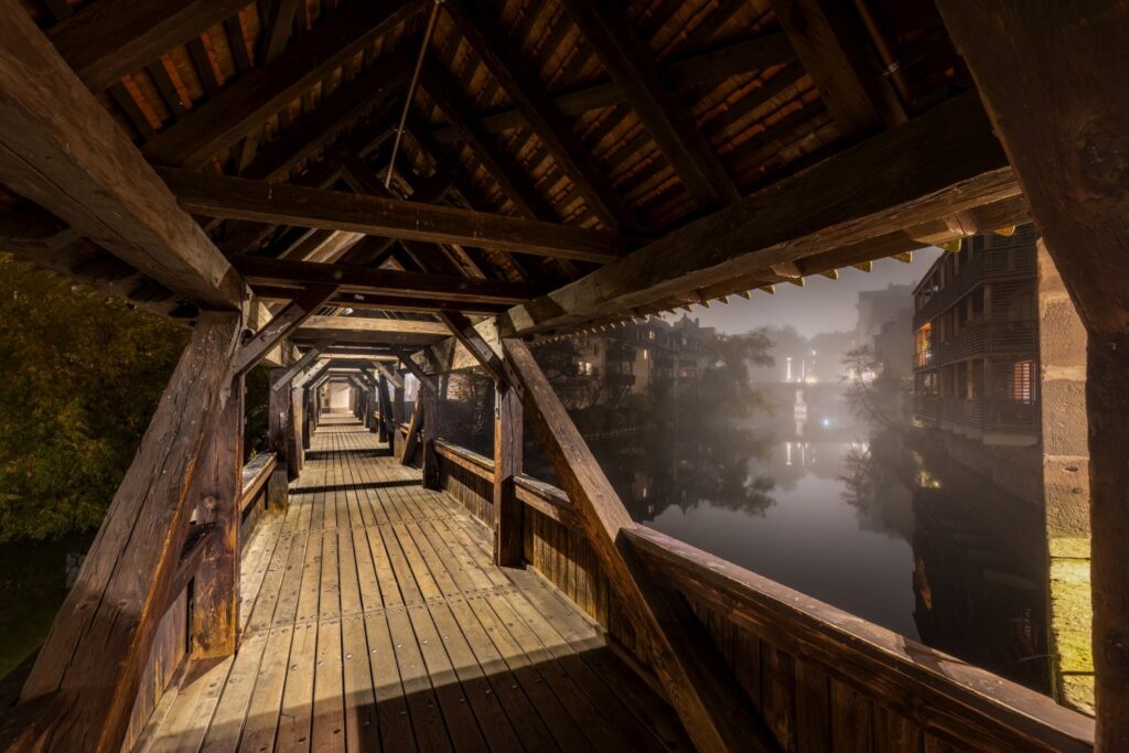 Das Bild zeigt einen langen, beleuchteten Holzdurchgang, von dem aus man eine neblige Flusslandschaft mit Gebäuden und Lichtern bei Nacht sieht.