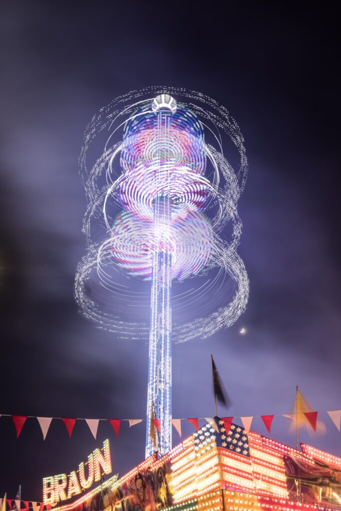 Nachtaufnahme eines beleuchteten Turms mit spiralförmigen Lichtspuren über einer bunten Kirmes.
