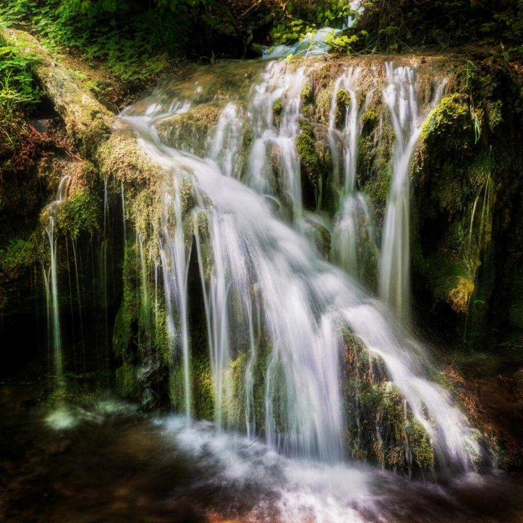 Wasserfall im Wald mit moosigen Felsstufen und sanften Kaskaden, grüne Naturkulisse.