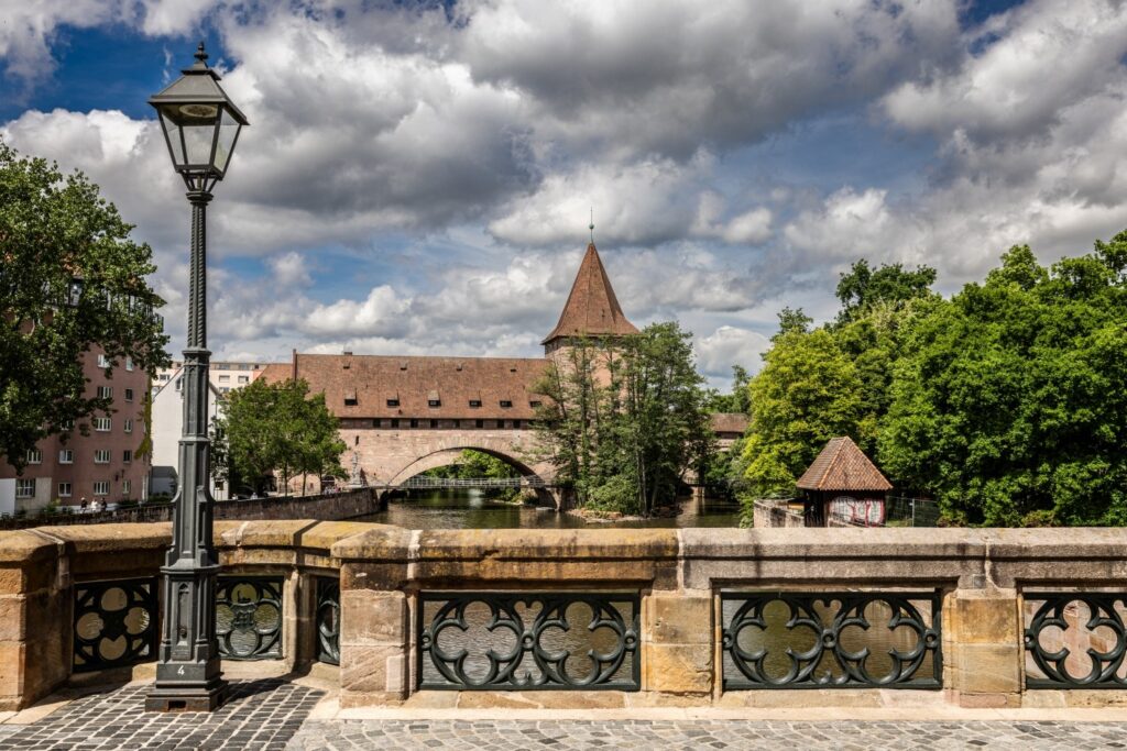 Im Vordergrund befindet sich ein alter Laternenpfahl auf einem steinernen Brückengeländer, das den Blick auf einen Fluss freigibt, der unter einer Bogenbrücke und entlang historischer Gebäude, darunter eines mit einem spitzen Turm, sowie vielen grünen Bäumen fließt, alles unter einem bewölkten Himmel.