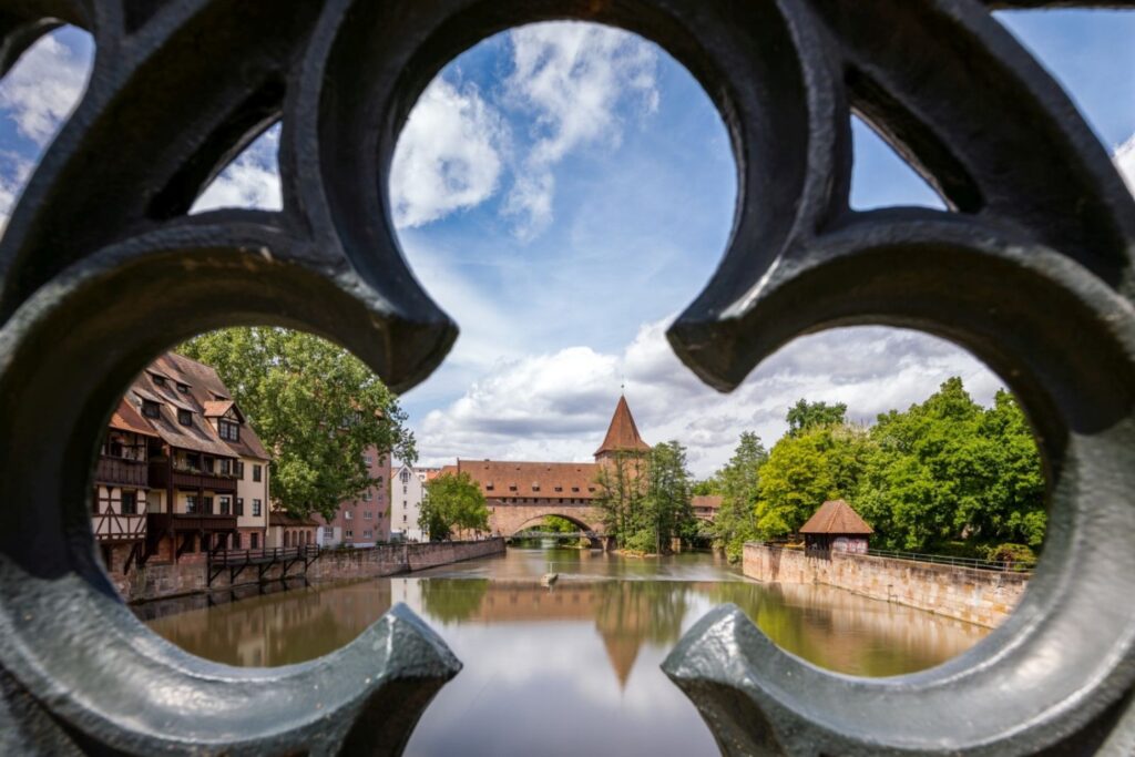 Durch ein dunkles, ornamentales Gitter blickt man auf einen Fluss, der von historischen Fachwerkhäusern, einer Brücke mit einem markanten Turm und grünen Bäumen unter einem bewölkten Himmel gesäumt wird, wobei sich die gesamte Szenerie im Wasser spiegelt.