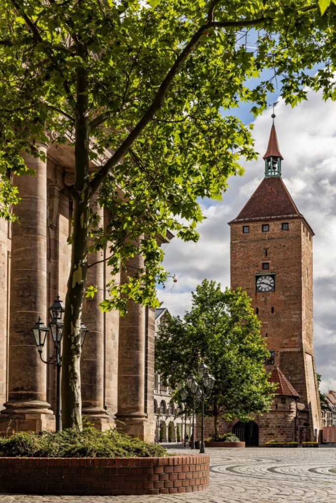 Das Bild zeigt einen gepflasterten Platz mit einem großen belaubten Baum im Vordergrund, flankiert von steinernen Säulen links und einem Backsteinturm mit Uhr und rotdachigem Turmhelm rechts unter bewölktem Himmel.