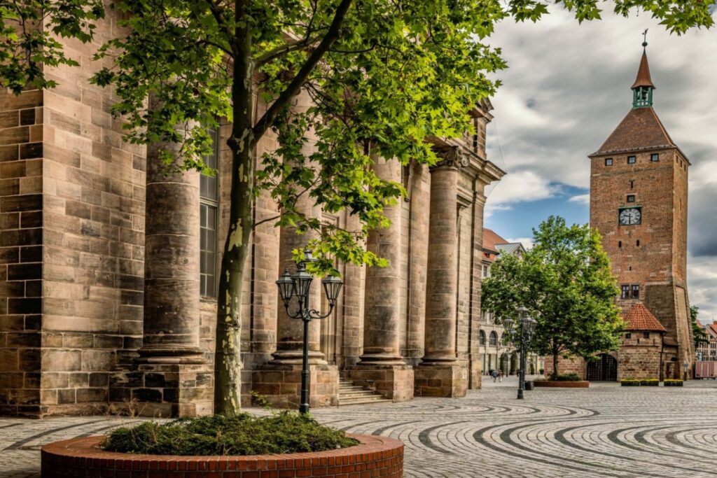 Auf einem gepflasterten Platz mit einem Baum und einer Laterne stehen ein historisches Steingebäude mit Säulen und ein hoher Backsteinturm mit Uhr unter einem bewölkten Himmel.