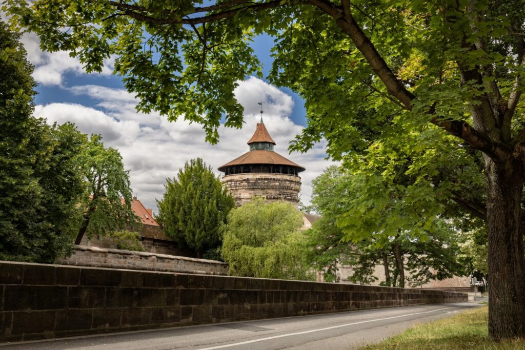 Ein runder Turm mit markantem braunem Dach ragt zwischen grünen Bäumen und über einer langen Steinmauer hervor, vor der eine Straße unter einem bewölkten Himmel verläuft.