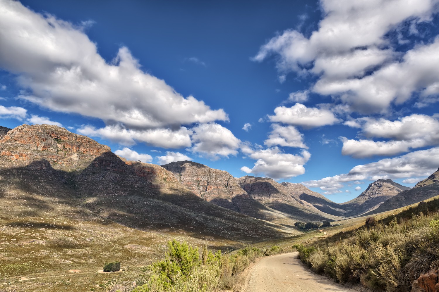 Berglandschaft im Tal, Schotterweg schlängelt sich durch Gräser; blauer Himmel mit weißen Wolken.