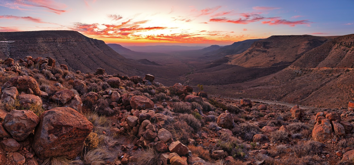 Sonnenuntergang über einer kargen Wüstenlandschaft mit tiefem Canyon, rotem Fels, Kies und Sträuchern im Vordergrund.