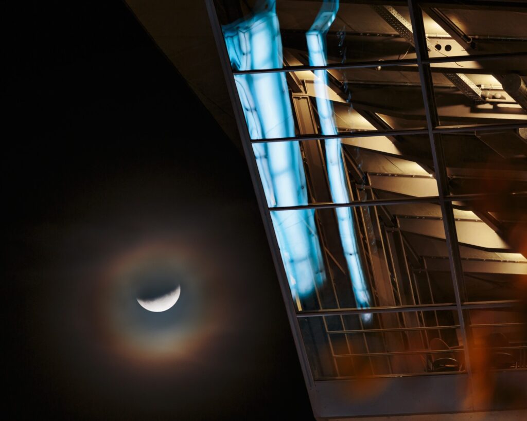 Nachtaufnahme eines modernen Glasgebäudes mit blauen Lichtern; Mond mit Halo-Effekt im dunklen Himmel.