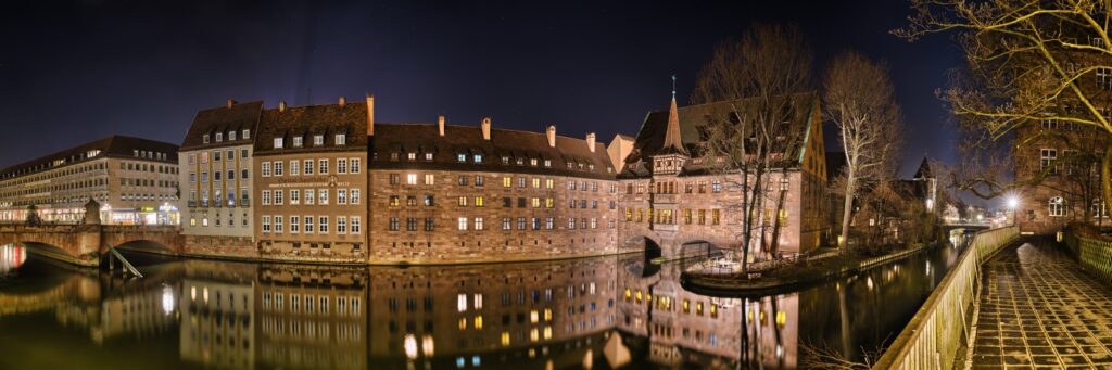Nächtliches Panoramabild einer historischen Stadt am Fluss, beleuchtete Fassaden, Brücke, Uferweg und Reflektionen im Wasser.