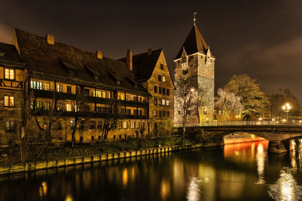 Eine nächtliche Szene zeigt eine historische Flusslandschaft mit beleuchteten Gebäuden, einem markanten Turm und einer Bogenbrücke, deren Lichter sich im ruhigen Wasser spiegeln.