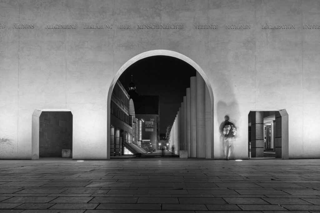 Nachtaufnahme eines monumentalen Betonportals mit rundem Bogen und Durchgang; UN-Menschenrechte-Inschrift an der Wand.