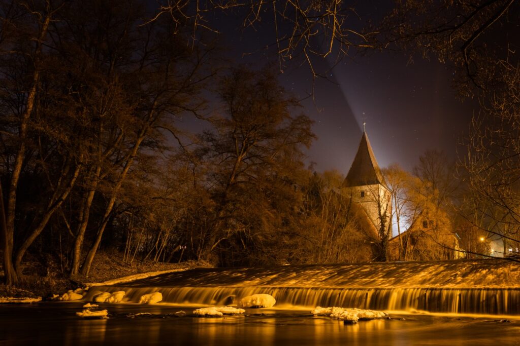 Nachtaufnahme eines historischen Turms mit Spitzdach, beleuchteter Wasserfall und von Bäumen gesäumtem Flussufer.