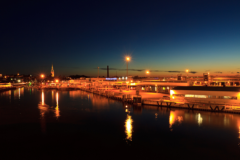 Hafen bei Abenddämmerung mit beleuchtetem Pier, Kirchturm und glitzernden Wasserreflexionen.