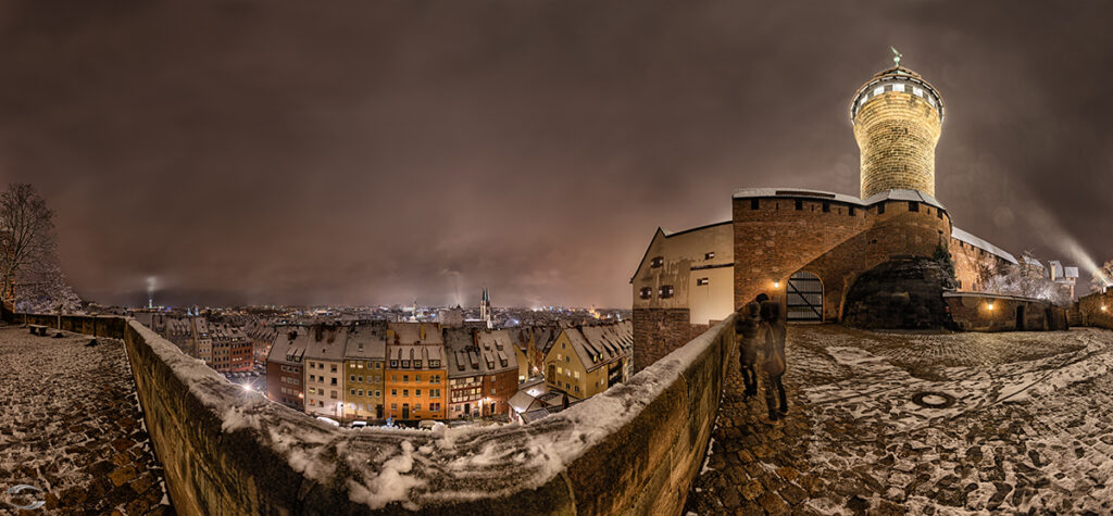Nachtpanorama einer historischen Burg mit Rundturm, verschneiter Stadtmauer und beleuchteter Altstadt.