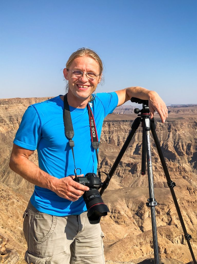 Ein Fotograf in einem blauen T-Shirt steht an einem Aussichtspunkt in einer Wüstenlandschaft, hält eine Kamera in der Hand und hat ein Stativ neben sich. Die beeindruckenden Felsen und die weite Landschaft sind im Hintergrund sichtbar.