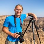 Ein Fotograf in einem blauen T-Shirt steht an einem Aussichtspunkt in einer Wüstenlandschaft, hält eine Kamera in der Hand und hat ein Stativ neben sich. Die beeindruckenden Felsen und die weite Landschaft sind im Hintergrund sichtbar.