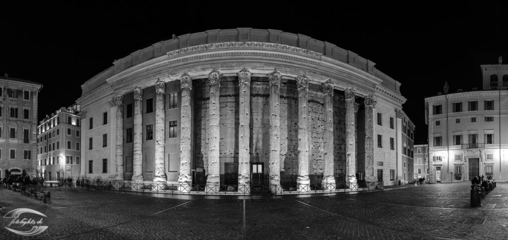 Nachtaufnahme eines antiken Tempelbaus mit dorischen Säulenfronten auf einem historischen Stadtplatz.