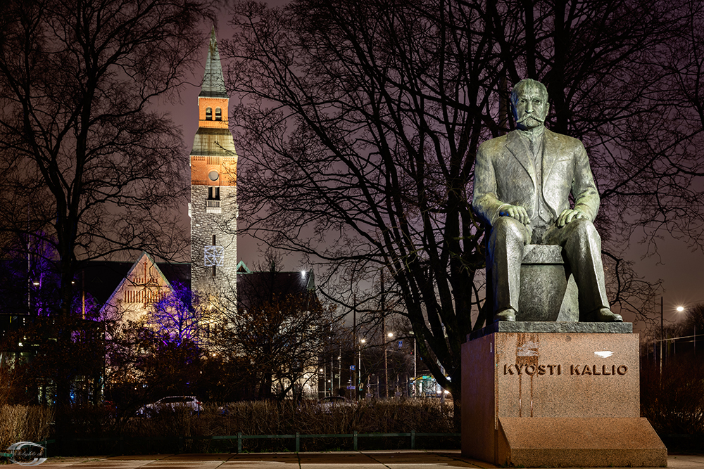 Nachtaufnahme einer sitzenden Statue eines finnischen Politikers vor beleuchtetem Kirchturm und Bäumen im Stadtzentrum.